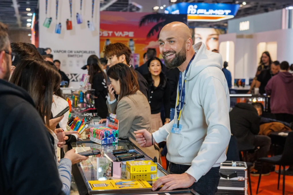 Un exposant souriant présentant du matériel et des e-liquides aux visiteurs sur son stand au salon Vapexpo Paris